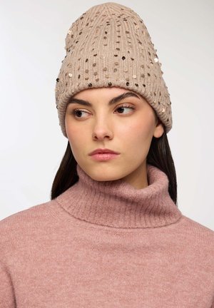 Beanie beige lavorato a maglia con paillettes decorative, dal design morbido. La texture è soffice, con motivi a coste. Indossato con un dolcevita rosa.