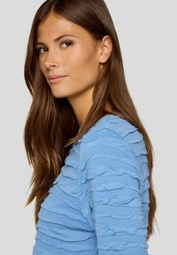 Hellblauer, strukturierter Oberteil mit horizontalen Rüschen, aus weichem Material und in taillierter Form. Glatter Ausschnitt und lange Ärmel.