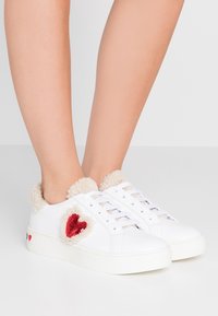 Vita sneakers med en fluffig beige krage, med en detalj i form av ett rött paljett-hjärta och en texturerad vit gummisula.