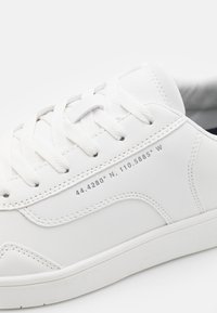 White leather sneaker with white laces, decorative stitching, and printed coordinates "44.4280° N, 110.5885° W" on the side panel.