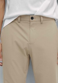 Gros plan sur une personne portant un pantalon beige avec un bouton noir et un t-shirt blanc rentré.