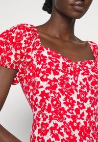 Rotes Blumenkleid mit weißen Mustern, quadratischem Ausschnitt und Puffärmeln. Das Material wirkt leicht und hat eine glatte Textur.