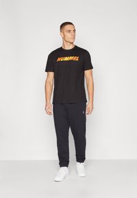 Juodas trumpomis rankovėmis marškinėliai su oranžiniu "HUMMEL" logotipu, derinami su tamsiomis sportinėmis kelnėmis ir baltais sportiniais batais, aiškus fonas.