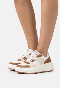 Białe i brązowe sneakersy z teksturowanym wzorem, z zamszowymi akcentami, okrągłym noskiem, grubą żłobioną podeszwą i kontrastującą fioletową pętelką na pięcie.