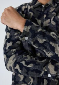 Persoon in een marineblauwe en grijze camouflage lange mouwen jas met drukknopen bij de manchetten en een rits aan de voorkant, met de armen gekruist op de borst.