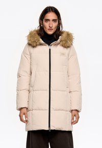 Abrigo acolchado beige con cremallera negra, gran capucha forrada de pelo y dos bolsillos laterales. Presenta una textura acolchada y un parche de logo.