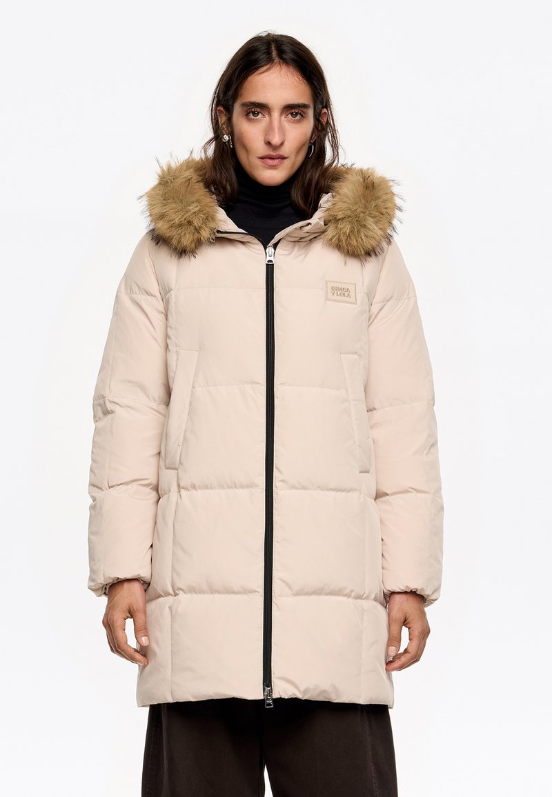 Abrigo acolchado beige con cremallera negra, gran capucha forrada de pelo y dos bolsillos laterales. Presenta una textura acolchada y un parche de logo.