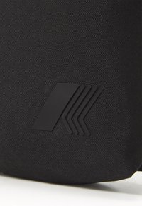 Schwarze Stofftasche mit strukturierter Oberfläche und mattem schwarzen Logo mit kantigen Linien. Schlichtes, funktionales Design ohne zusätzliche Verzierungen.