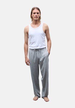Jeune homme aux longs cheveux portant un t-shirt blanc sans manches et un pantalon de survêtement gris, debout pieds nus avec une main dans la poche.