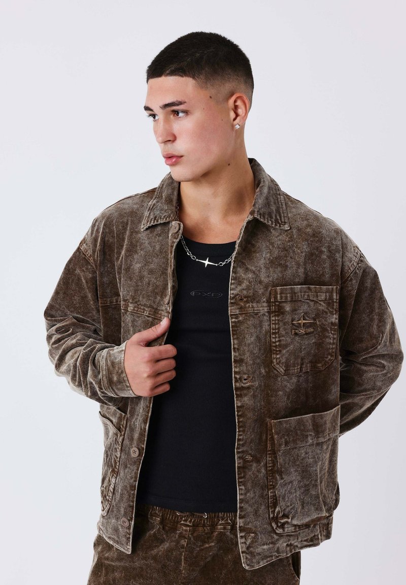Jeune homme portant une veste et un pantalon en velours côtelé marron, avec une chemise noire et un collier étoile argentée, tourné vers la gauche sur un fond uni.