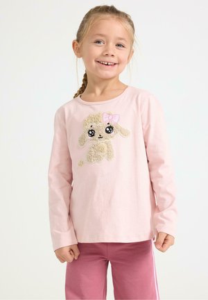 Smilende ung pige med flettet hår iført en lyserød langærmet skjorte med et tekstureret hvalpedesign og matchende lyserøde bukser.