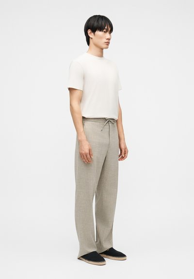 Άνδρας που στέκεται πλάγια, φοράει απλό λευκό κοντομάνικο πουκάμισο, μπεζ παντελόνι με κορδόνι και μαύρα παπούτσια χωρίς κορδόνια με ανοιχτόχρωμη σόλα.