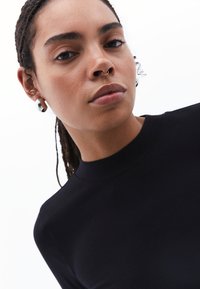 Top nero a collo alto con maniche lunghe, texture liscia e design aderente. La modella indossa un grande orecchino circolare e un anello al setto.