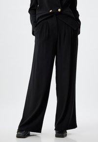 Pantalons larges noirs en tissu léger avec des plis, dotés de poches latérales et d'une coupe décontractée, associés à des chaussures texturées noires.