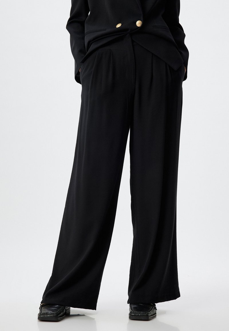 Pantalons larges noirs en tissu léger avec des plis, dotés de poches latérales et d'une coupe décontractée, associés à des chaussures texturées noires.