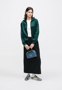 Green velour zip jacket, white top, black long skirt, and gray soccer shoes. Holding blue handbag with embossed logo.