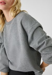 Pull en maille gris avec un col ras du cou, des épaules tombantes et des manches retournées, présentant une finition douce et texturée.