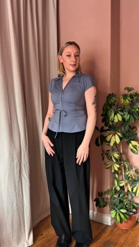 Γυναίκα με κοντά ξανθά μαλλιά, ντυμένη με εφαρμοστή γκρι μπλούζα και μαύρο παντελόνι με φαρδιά μπατζάκια, στέκεται κοντά σε πράσινο φυτό σε γλάστρα και μπεζ κουρτίνα.