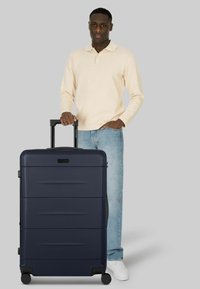 Hombre con camisa de manga larga color crema y jeans azules sosteniendo la asa de una maleta grande azul marino con ruedas sobre un fondo gris.