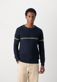 Marineblå genser i strikket stoff med strukturt mønster, med en horisontal stripe i svart og hvitt. Rund hals, lange ermer og ribbestrikkede mansjetter.