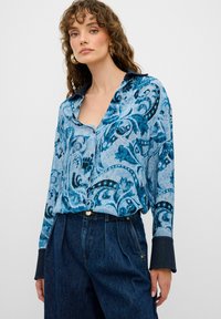 Camicetta a maniche lunghe in blu e turchese con motivo floreale paisley, caratterizzata da un colletto con intaglio e polsini blu scuro. Indossata con pantaloni di jeans a vita alta.