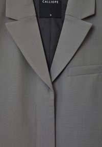 Blazer grigio con colletto a scenda, taschino sul petto singolo e texture liscia. L'interno presenta una fodera nera. Etichetta del marchio "CALLIOPE" visibile.