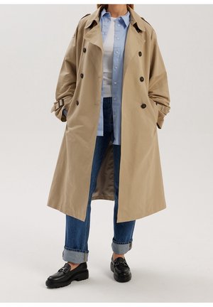 Person iført beige trenchcoat, lyseblå skjorte med knapper, hvid t-shirt, opkørte blå jeans og sorte loafers, stående med hænderne i lommerne.