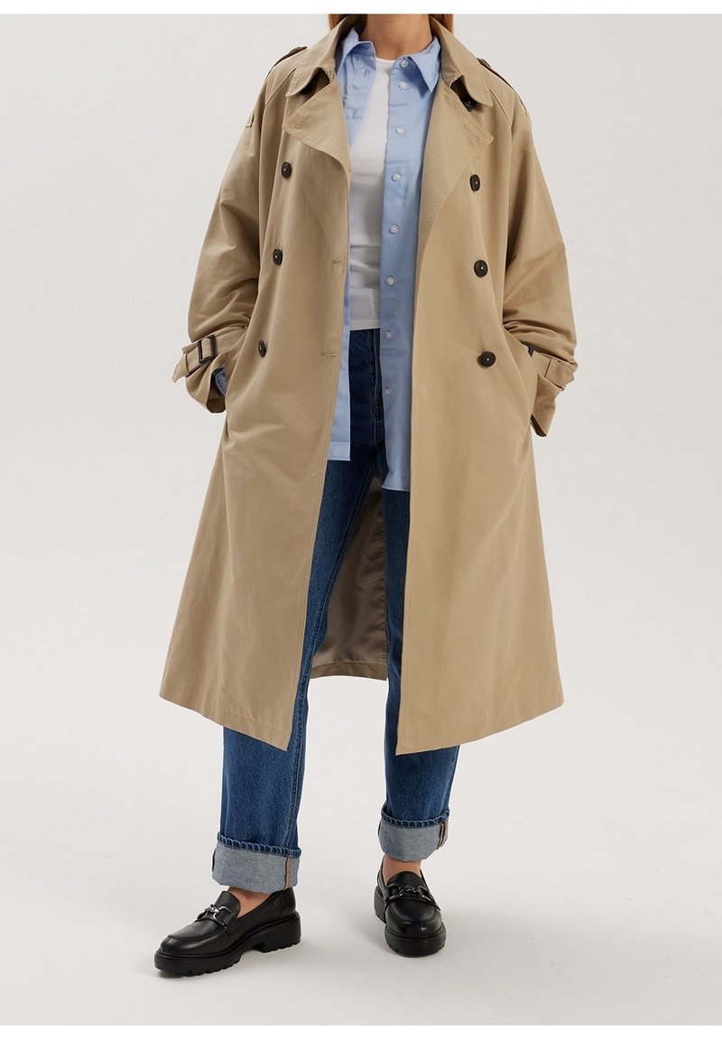 Personne portant un trench-coat beige, une chemise bleu clair à boutons, un t-shirt blanc, un jean bleu roulé et des mocassins noirs, debout les mains dans les poches.