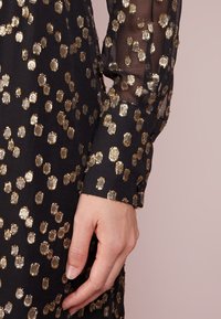 Robe en tissu noir avec un motif à pois dorés et des manches longues transparentes. Détail de poignet à boutons avec une texture fine dans le tissu.