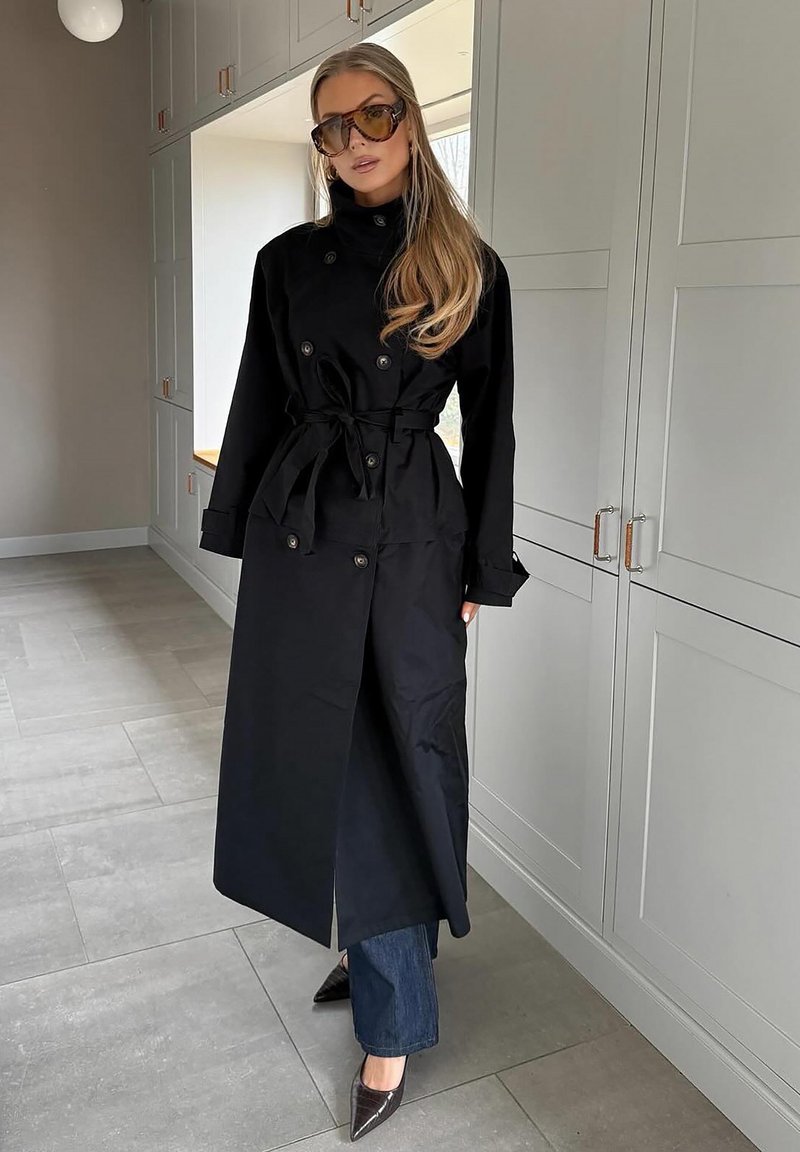 Čierny trenčkot s dvojradovým zapínaním a opaskom, skombinovaný s tmavými denimovými džínami a špicatými lodičkami, v modernom interiéri.
