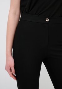 Zwarte, getailleerde broek met een gladde textuur, voorzien van een brede tailleband en een zilveren knoopaccent. Close-upweergave die de details van de stof toont.