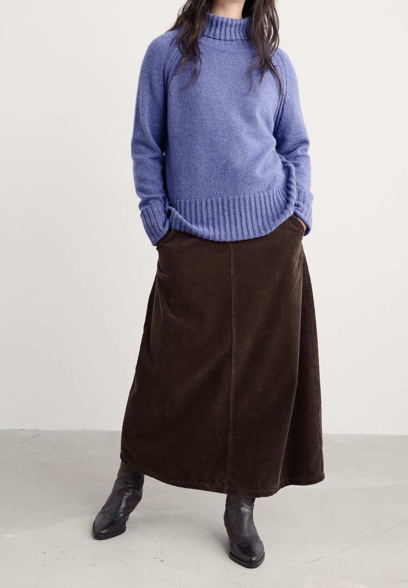 Femme portant un pull à col roulé bleu, une longue jupe marron en velours côtelé et des bottines noires, debout les mains dans les poches.