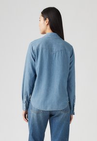 Lyseblå denimskjorte med lange ermer, spiss krage og knappecuff. Bakdelen har en yoke og en buet hem-design.