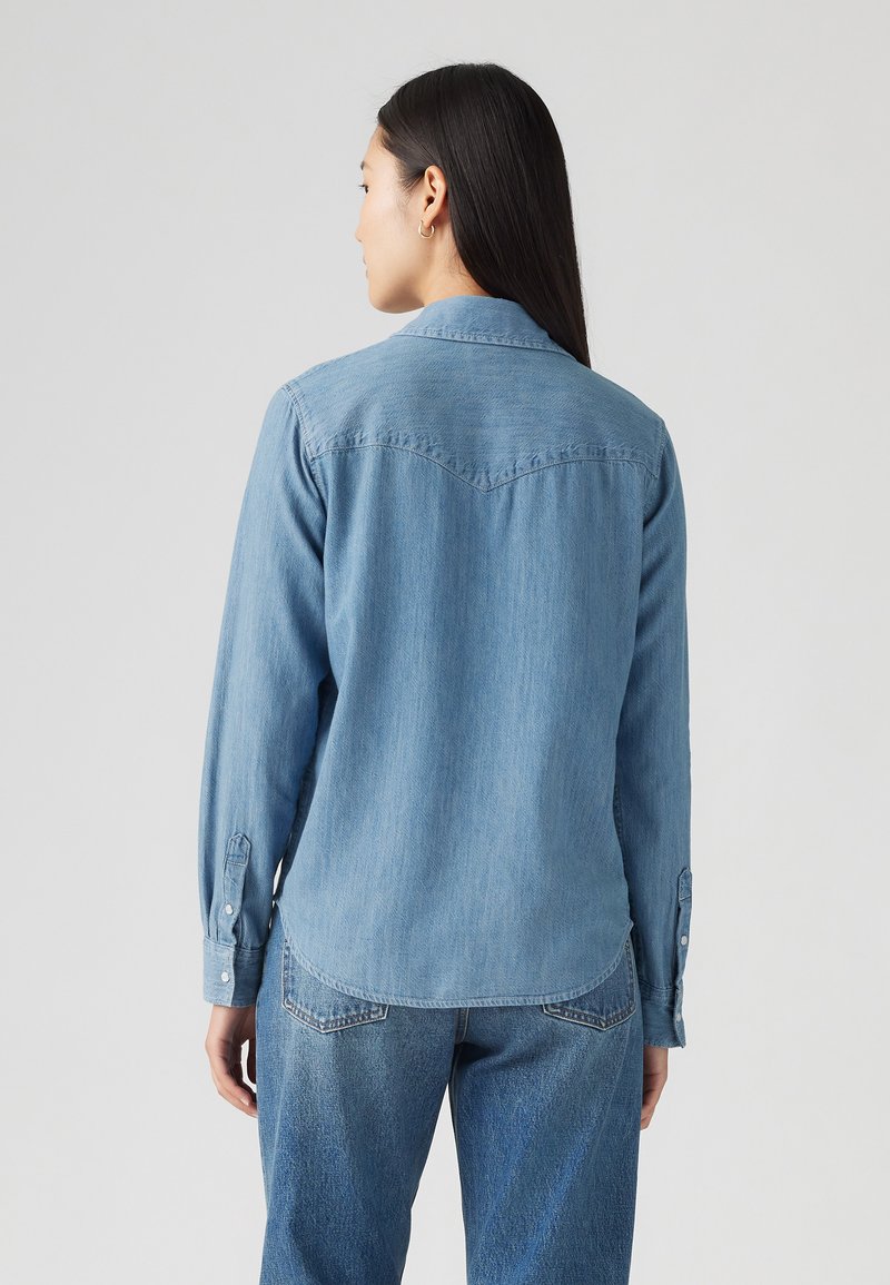 Chemise en denim bleu clair à manches longues, avec un col pointu et des poignets à boutons. Le dos présente un empiècement et un design d'ourlet courbé.