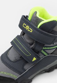 Zapato de senderismo de media altura en azul oscuro con malla gris, acentos y costuras en verde lima. Presenta dos correas de velcro y una suela de goma texturizada.