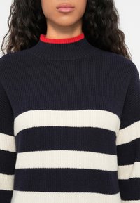 Stickad tröja med hög halsringning. Mörk marinblå bas med vita horisontella ränder och en röd ribbad krage. Texturerad, mjuk tyg.