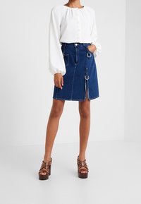 Gonna di denim con taglio dritto e spacchetto laterale, anelli metallici e dettaglio intrecciato, abbinata a una camicetta bianca con bottoni e sandali con tacco.
