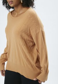 Lysebrunt oversized sweater med ribbede kanter ved hals og i bunden. Har detaljerede forhøjede mønstre på forsiden og lange ærmer.