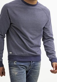 Sudadera azul marino con patrón texturizado, cuello redondo y puños acanalados. Combinada con vaqueros desgastados de lavado claro.