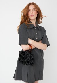 Črna perlična torbica z teksturirano površino in okroglimi ročaji leži v položaju prekriženih rok, v kombinaciji s sivo obleko s kratkimi rokavi in ovratnikom.