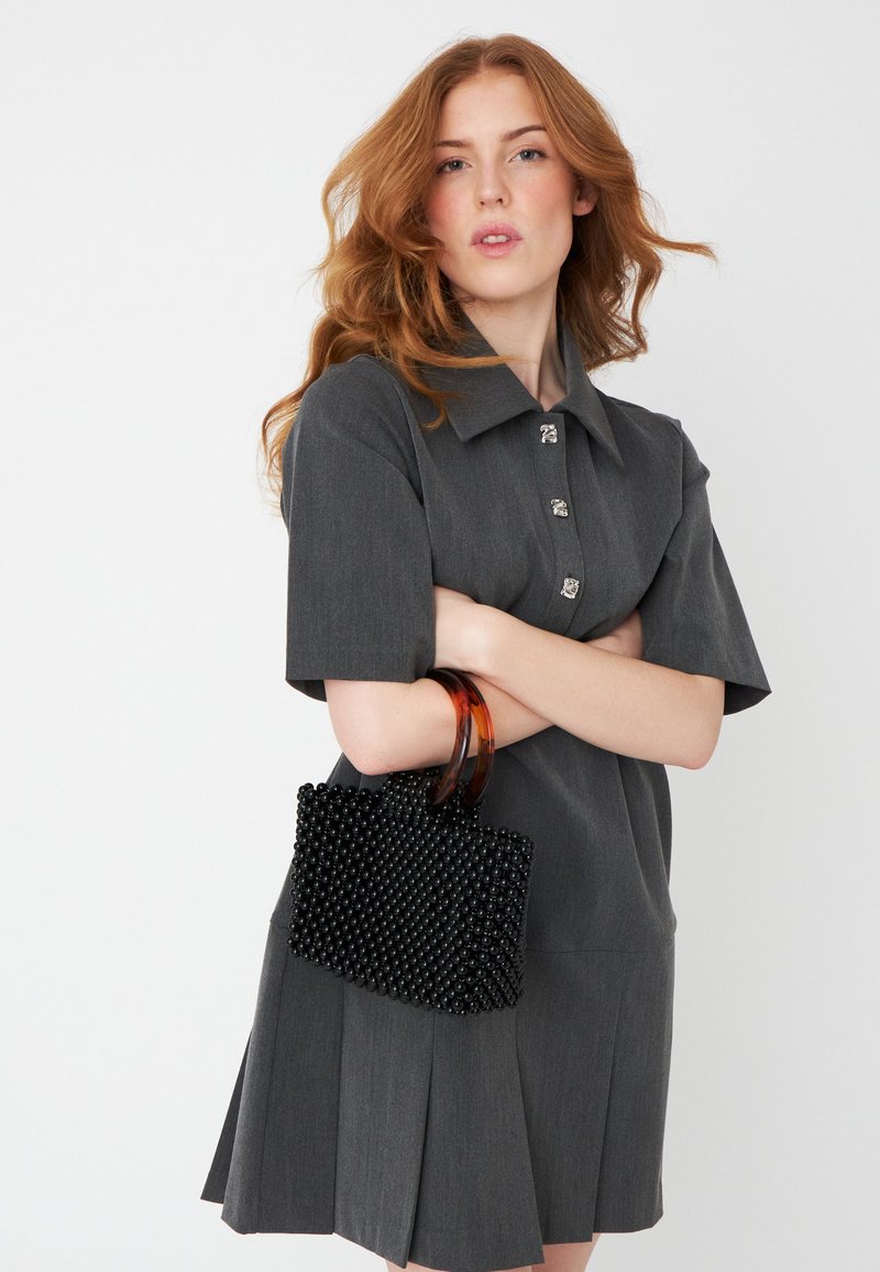 Črna perlična torbica z teksturirano površino in okroglimi ročaji leži v položaju prekriženih rok, v kombinaciji s sivo obleko s kratkimi rokavi in ovratnikom.