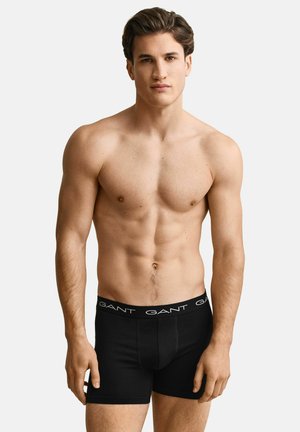 Junger muskulöser Mann mit kurzen Haaren, der schwarze GANT-Boxershorts trägt und vor einem schlichten hellen Hintergrund steht.