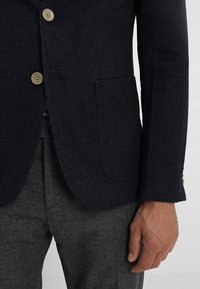 Close-up of a man's torso wearing a textured dark navy blazer with beige buttons and gray patterned pants, hand relaxed at side.