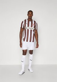 Nogometni dres z rjavimi in belimi črtami ter kratkimi rokavi, kombiniran z belimi hlačami in nogavicami. Na prsih ima logotip in blagovno znamko.