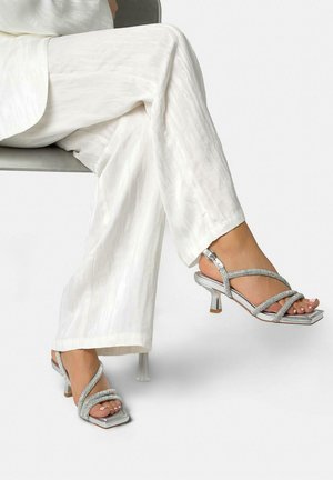 Piernas cruzadas con pantalones blancos y sandalias de tacón plateadas con tiras y punta cuadrada, sentada en una silla gris contra un fondo blanco.