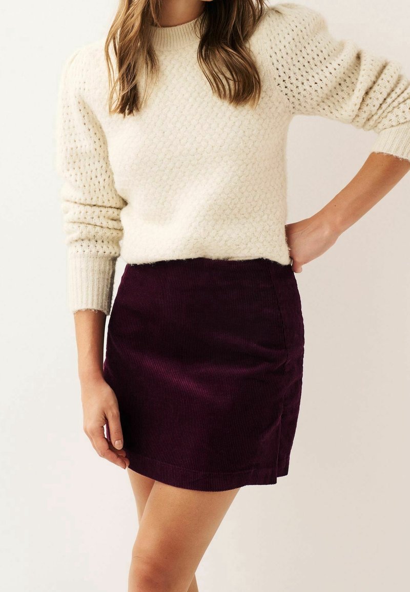 Cremfarbener Strickpullover mit strukturiertem Design, kombiniert mit einem bordeauxroten Cordrock in Mini-Länge mit glatter Oberfläche und seitlicher Nahtdetail.