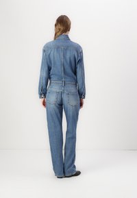 Τζιν ολόσωμη φόρμα σε ανοιχτό μπλε, με μακριά μανίκια, κουμπιά snap και ελασμένη μέση. Χαλαρή ίσια γραμμή με πίσω τσέπες.