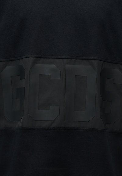 Schwarzer Stoffhintergrund mit großen, erhabenen, dunklen glänzenden Buchstaben „GCDS“, die teilweise in der Mitte sichtbar sind.