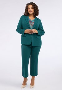 Donna con capelli ricci che indossa un giacca e pantaloni di colore teal, una blusa a fantasia e tacchi beige, in piedi davanti a uno sfondo bianco.