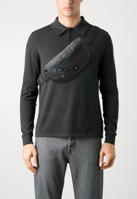 Borsa a tracolla grigia realizzata in tessuto imbottito, con chiusura a zip, patch con logo e tracolla regolabile, indossata sopra una camicia a maniche lunghe scura.